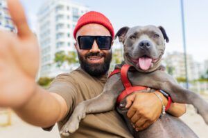 portrait of happy man in red hat and sunglasses with american terrier in dogs walking area park in City